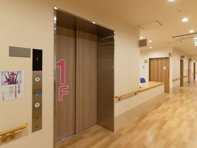 1階表示の上品なエレベーター エレベーター 手すりが設置された温かみのある通路があり、スタッフの方々が笑顔で対応する受付も見えますので、ご安心してお過ごしいただけます。
