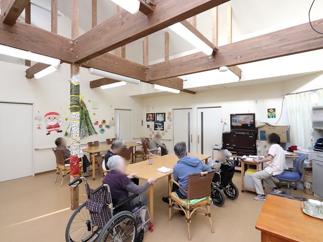 開放感ある共有空間 談話室 開放的な天井と暖かい木目の共有スペースで、利用されている方々が穏やかな時間を過ごしています。テレビを見ながらお互いの交流も深められ、ゆったりとした雰囲気が広がっています。