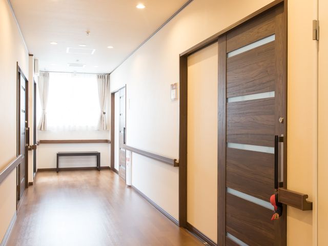 廊下の木目調のドア 廊下 木目調で落ち着いた色合いのドアが特徴の通路で、訪れる皆さまの心地よい移動をサポートします。明るい窓辺にはベンチも設けられており、休憩もできる配慮が見受けられます。