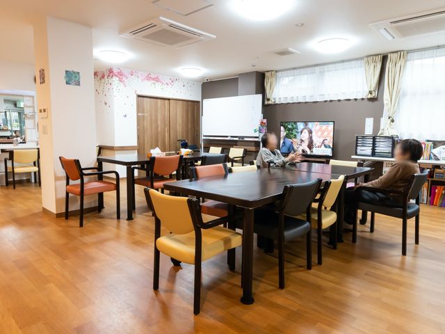 共有スペースでくつろぐ人々 共有スペース 広々とした食堂で、利用者の方々がテレビを楽しみながらくつろいでいます。ゆったりとした時間が流れる環境で、働いてみませんか。