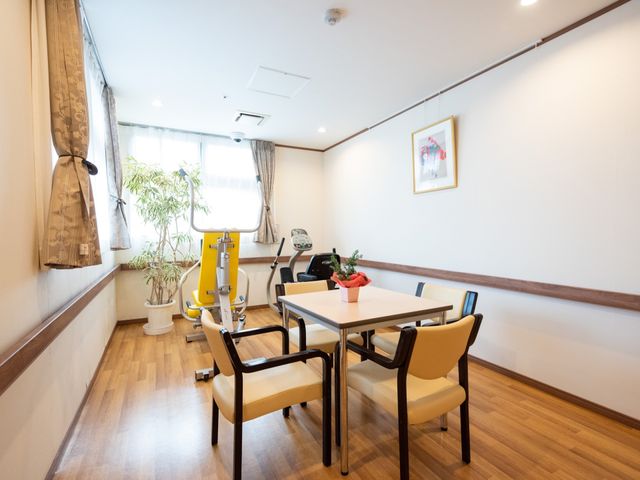 機能訓練器具とテーブルの部屋 部屋 広々とした部屋には機能訓練用の器具とテーブルが整備され、心身の活動をサポートしています。窓からの自然光が明るい雰囲気を演出しており、快適な空間で過ごせます。