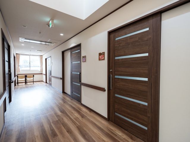 明るく清潔な廊下 通路 床が木目調で落ち着いた雰囲気の廊下は、スタッフと入居者の円滑な移動を支えます。明るい窓辺にはベンチも設けられ、安らぎのひとときを過ごせる空間です。