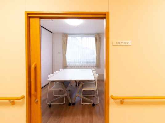 地域交流室と書かれた看板がある小部屋。カーテンの掛かった窓があり、白い大きなテーブルと椅子が置かれている。
