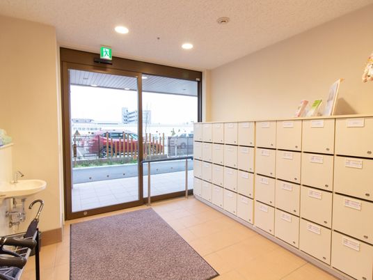 ガラス張りの自動ドアがある明るい玄関。壁際には扉のついた白い大きな下駄箱がある。足元には玄関マットが敷かれている。
