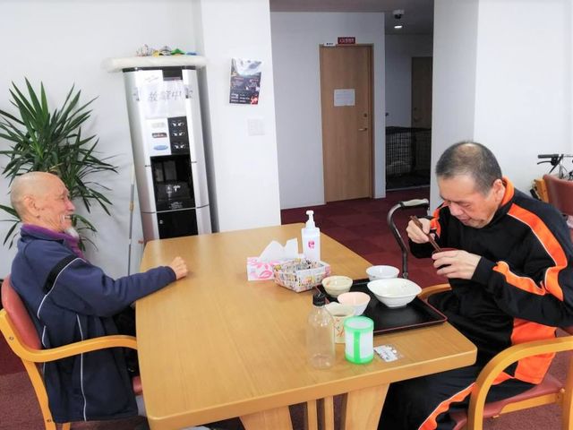 食事を楽しむ高齢者二人 食堂 笑顔で会話しながら一緒に食事ができる、アットホームな雰囲気のダイニングです。明るい部屋でリラックスした時間を過ごしていただけます。