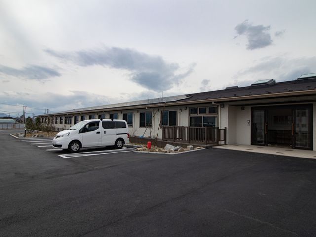 低層建築の外観 駐車場 広々とした駐車場と木製の手すりが設置されたモダンな建物外観です。来訪者も利用者も明るい雰囲気で迎えられます。
