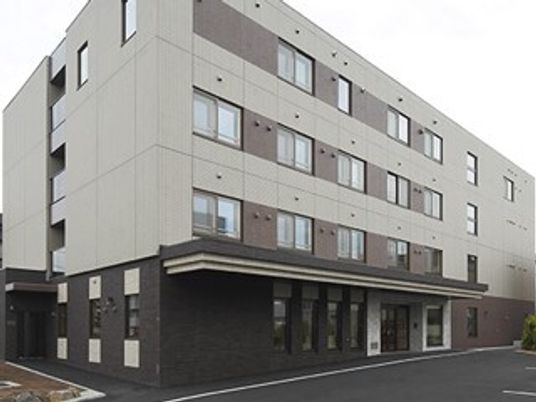 施設の写真 4階建ての建物は1階部分がダークブラウン、2階以上は白でたくさんの窓が付き、玄関周りに段差はなく、車寄せが付いている。