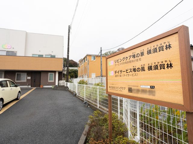 施設の看板と外観 駐車場 訪れる方を温かく迎える木製の案内板があり、静かな住宅街に調和する落ち着いた色合いの建物が見えます。