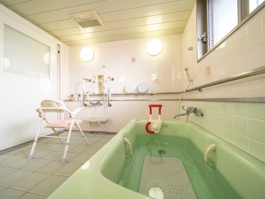 窓がある広々とした浴室に黄緑色の浴槽が設置されている。シャワーの前に、背もたれと肘掛けが付いた椅子が置いてある。