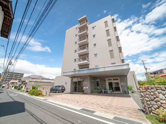 地上７階建ての施設の前に自動車が１台駐車してある。道路と敷地との間に段差が少なく、間口も広くなっている。