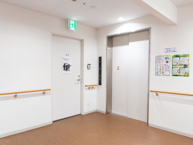 清潔な施設の廊下 エレベーター 広々とした廊下は清潔感があり、壁には利用者の安全に関する掲示物もございます。利用者の方々が安心して日々を過ごせるよう、細やかな配慮が施されています。