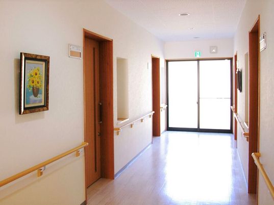 施設の写真 ヒマワリの絵画が飾られた廊下はまっすぐで、両サイドには手すりが付き、居室のドアが並んでいる。突き当りには大きなガラス戸があり、日当たり良好。