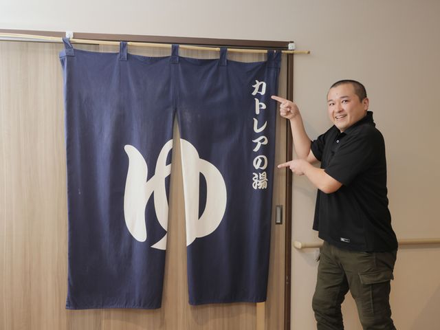 男性が示す浴室暖簾 浴室 浴室の入り口には、大きな暖簾が掛かっており、利用者を優しく迎え入れます。