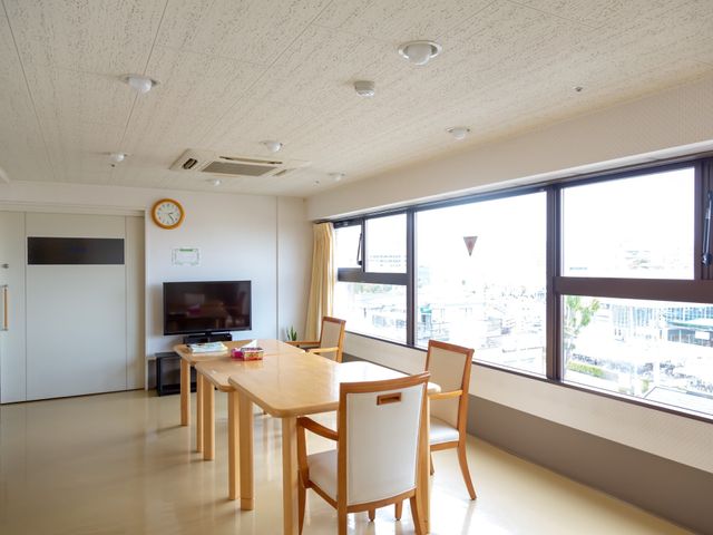 明るい居室のテーブルとテレビ 共有スペース ゆったりと配置されたテーブルと椅子があり、テレビも完備された休憩室です。大きな窓からは自然光がたっぷりと差し込みます。