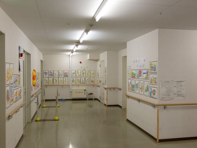 廊下に飾り付けられた壁 共用廊下 壁にはカラフルな作品が飾られ、歩行をサポートする手すりが整備された温もりのある空間です。安心して日々を過ごせる環境で、新たな職場を考えるのにふさわしい場所と言えます。