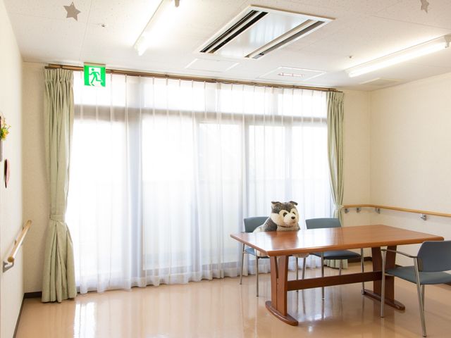 明るい居室の一角 多目的室  ゆったりとしたテーブルと椅子が配置され、窓から自然光が差し込む、暖かみのある休憩スペースです。柔らかな色合いの壁とカーテンが心地よい雰囲気を醸し出しています。