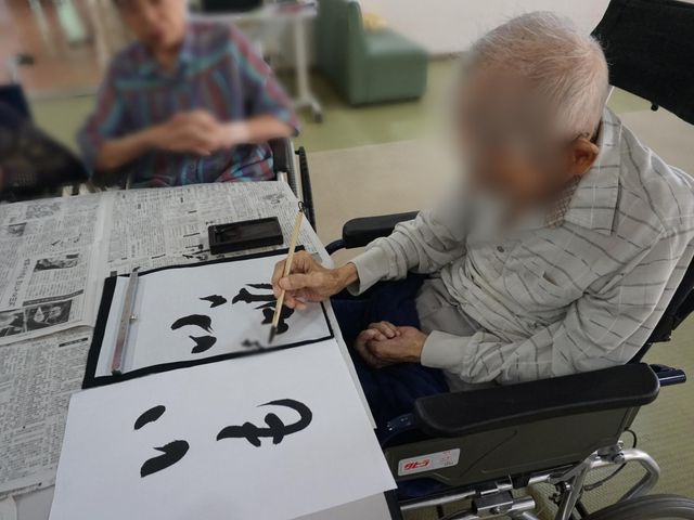 書道する人々の居室 居室 車椅子の方がリラックスした雰囲気で書道を楽しんでいます。明るい空間での趣味の時間です。