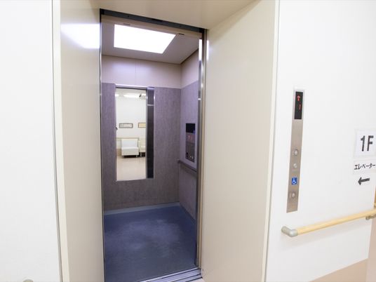 長方形の鏡が、エレベーター内部の壁に設置されている。右側の壁にはボタンパネルがあり、その下に手すりが付いている。