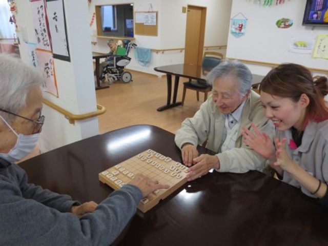 将棋を楽しむ居室の様子 趣味の時間 ケアスタッフが笑顔で対局を見守る、温もりある暮らしの一コマです。交流が生まれる居心地の良いスペースがあります。