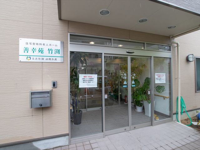 エントランスの扉と看板 入口 ガラス扉の先には観葉植物が置かれ、明るく開放的な雰囲気のエントランスです。安心感をもたらすような、きれいに整えられた入口がお出迎えいたします。