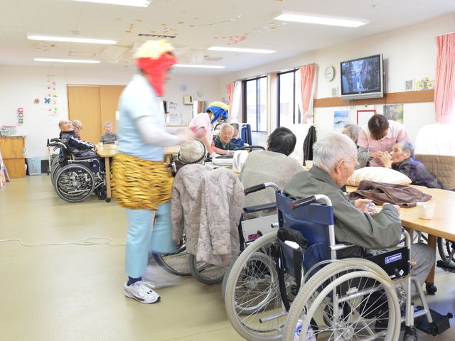 活動的な居室の様子 談話室 明るく広々とした居室で、スタッフと利用者が楽しげに過ごしており、笑顔があふれている光景です。動きやすい服装で、元気な雰囲気が伝わります。