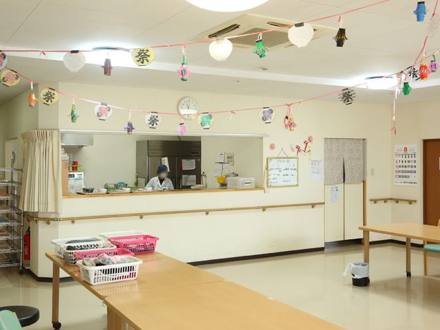 明るい共有スペースの風景 食堂 季節の飾りつけが施された共有スペースでは、利用者の皆さんが日々の食事や交流を楽しまれています。スタッフの方が配膳エリアで丁寧に準備を進めている様子がうかがえます。