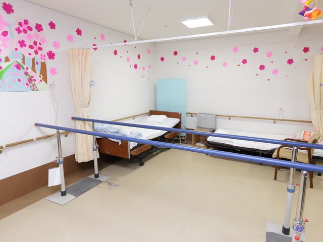 機能訓練室のベッド 訓練室 明るい色彩の壁紙が目を引く訓練室です。広々としたスペースには、リハビリ用のベッドと歩行訓練のための手すりが備えられ、快適な環境が整っています。