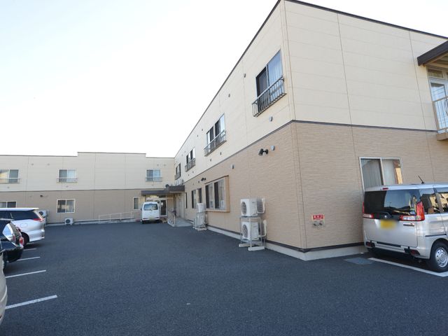 広々とした施設の駐車場 外観(駐車場側) 広々とした明るい駐車場が配置され、日々の送迎も安心して行える環境です。ゆとりを持って就業できる施設の一角をご覧ください。