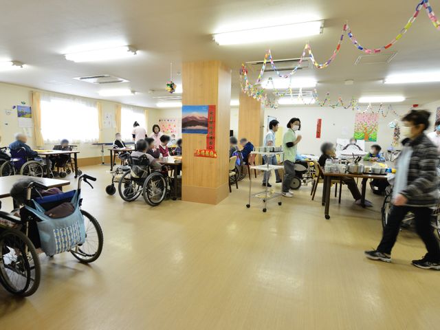 活気ある共用スペース 共有リビング 活動に満ちた広々としたエリアです。利用者が食事を楽しむ姿が見られ、スタッフも手厚いサポートを行っている様子が伺えます。