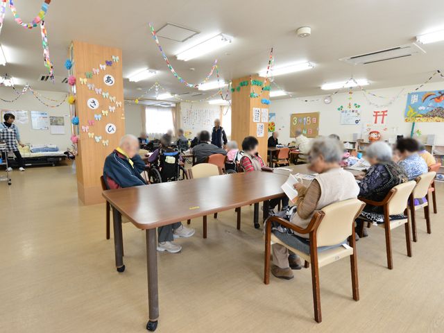 多世代が集う活動室 食堂と談話室 装飾が施されたアットホームな空間で、利用者さまがゆったりとお食事や憩いの時間を過ごされています。活動や交流を楽しむ姿が印象的です。