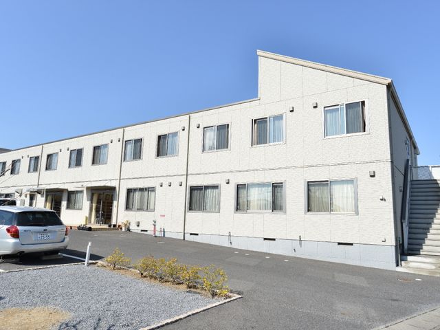 白壁の建物と駐車場 施設の外観 ゆったりとした駐車スペースが確保された明るい外観です。エントランスへのアクセスも良く、働く環境として安心感があります。