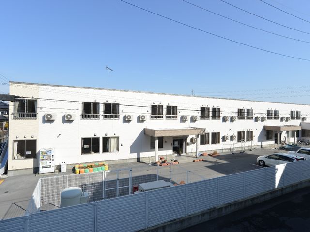 白壁の建物とエアコン 外観 広々とした駐車場がある清潔感のある施設です。明るい陽光が差し込む、快適な環境での就業が考えられます。