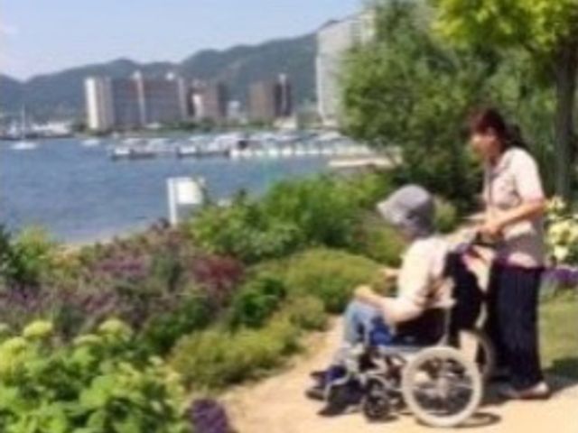 車椅子の利用者とスタッフ中庭 レクリエーション 広々とした中庭で、スタッフが車椅子の方をゆっくりとお連れしています。新緑の季節には、爽やかな風を感じながら、心地よい時間を過ごせるでしょう。
