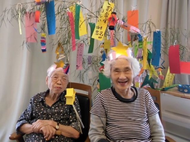 笑顔のお二人と七夕飾り イベント 七夕の願い事を書いた短冊が飾られた部屋で、明るく笑顔を見せているご利用者様がおり、楽しい雰囲気が感じられます。