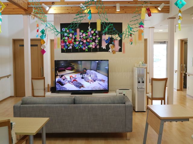 明るいリビングの様子 共有スペース ソファ前の大型テレビが映す番組に、訪れる人々が心温まるひと時を過ごせそうです。木の温もりあふれる装飾が施された共有スペースは、くつろぎの空間を提供しています。