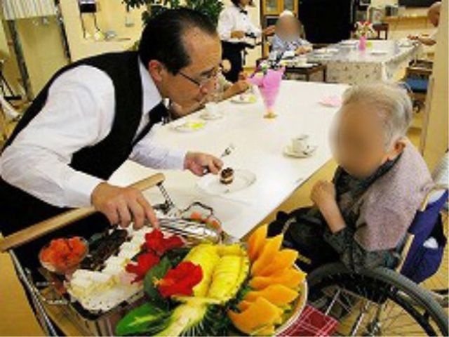 車椅子の利用者とスタッフ 日常 ケアスタッフが手作りのスイーツを提供する和やかな食堂の一コマです。利用者は笑顔で対話を楽しんでいます。