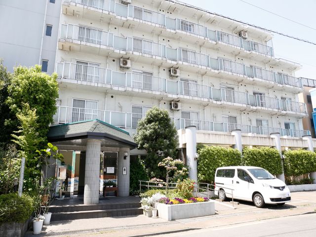 白い建物の明るいエントランス 外観 落ち着いた雰囲気のエントランスが迎える、白い建物の門前です。花々が彩りを添え、訪れる方々に安らぎを提供しています。