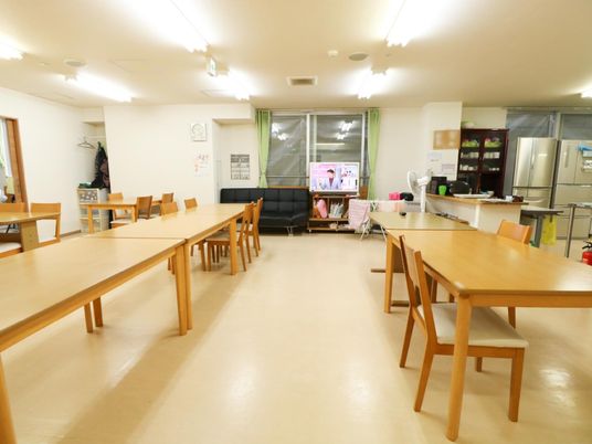 木製のテーブルや椅子が配置されており、窓側には液晶テレビやソファが置いてある。右側に複数の冷蔵庫や食器棚が並んでいる。