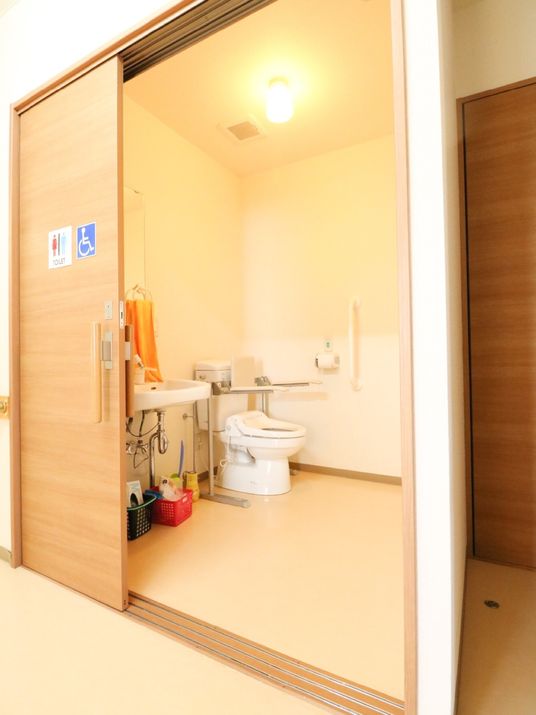 広々としたトイレ空間に、洋式便器と手洗器が設置されている。出入り口にトイレと車椅子のマークが貼ってある。