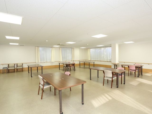 広々とした明るい多目的室 リビング ゆったりと配置された机と椅子があり、明るく開放的な空間です。ここでは様々な活動が行われ、コミュニケーションを深める場となっています。