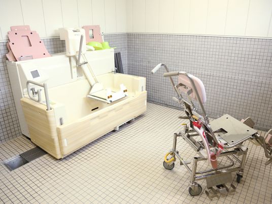 白色とグレイ色のタイル調仕様の浴室で、壁際には機械浴が設置されている。側面が半分を開ける浴槽になっている。