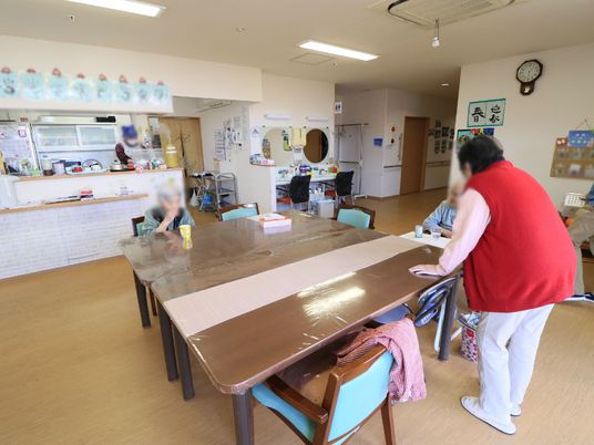 食堂では、テーブルが向かい合わせに配置され、入居者様皆様で向き合っている。キッチンでは、女性スタッフが食事の用意をしている。