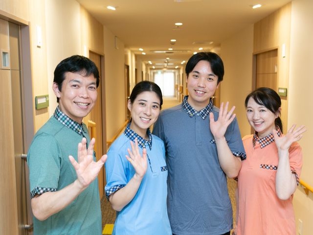 スタッフが並ぶ廊下 廊下   明るい廊下でスタッフの皆さんが笑顔で手を振っています。働く雰囲気のよさが伝わり、協力しやすそうな職場です。