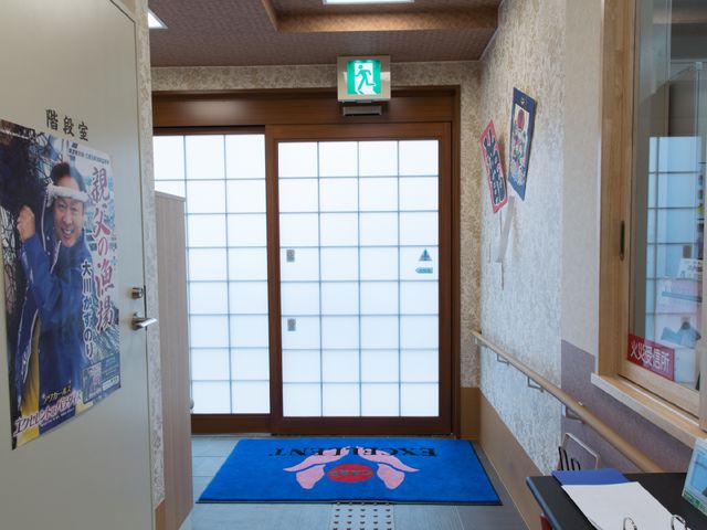 エントランスの明るい通路 施設玄関 暖かみのある光が差し込むエントランスは訪れる人々を温かく迎え入れます。壁には掲示物があり、賑わいを感じさせる空間です。