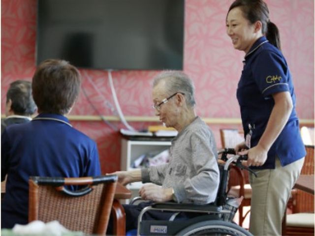 笑顔のスタッフと利用者様 ケア スタッフの方が利用者様を車椅子でサポートしており、温かな雰囲気の共有スペースでのさまざまなコミュニケーションが見受けられます。