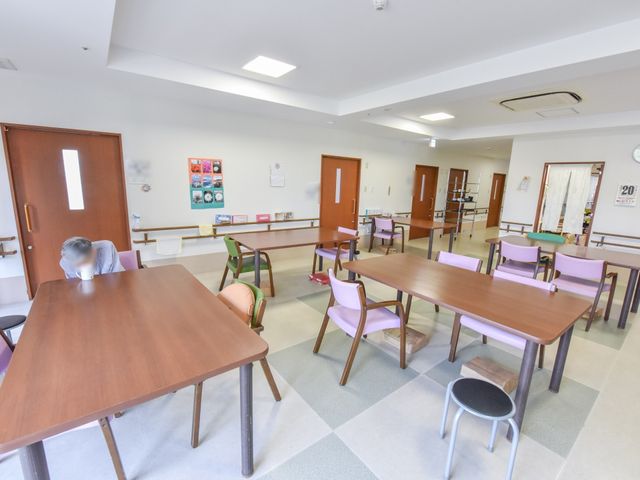 広々とした明るい食堂 リビングルーム 座席とテーブルが整然と配置された食堂で、心地よい空間です。職員の方と利用者様が談笑しながら心温まるひとときを過ごしている様子が伺えます。