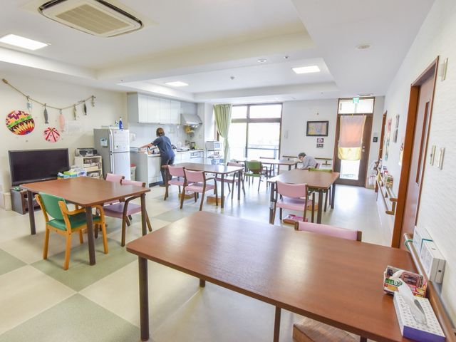 ゆったりした共有スペース 共有スペース 広々とした食堂には、テーブルが並び、キッチンで調理するスタッフの姿も見えます。窓から自然光が差し込む明るい環境で、利用者同士が交流を深める場となっています。