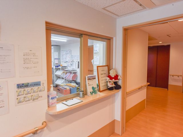 窓越しの明るい事務室風景 受付 窓越しに見えるスタッフルームは、整理整頓された作業スペースが確保されております。手厚いサポート体制が備わっています。