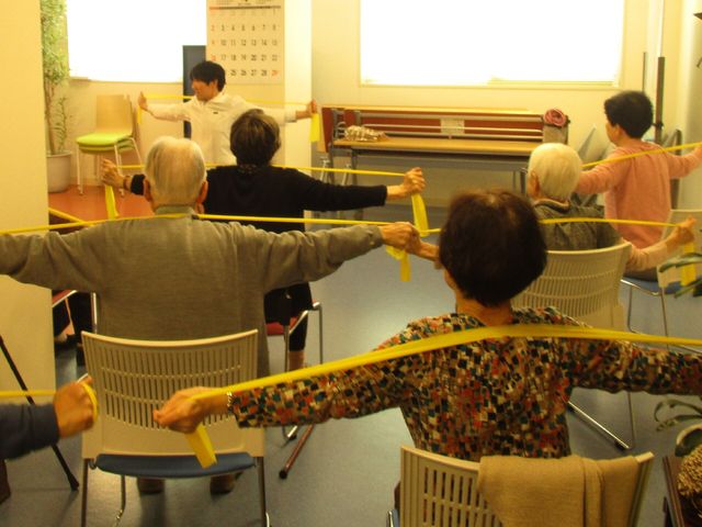 運動する人々の機能訓練室 体操教室 明るい機能訓練室で、スタッフの指導のもと、利用者様が健康維持のための体操をされています。