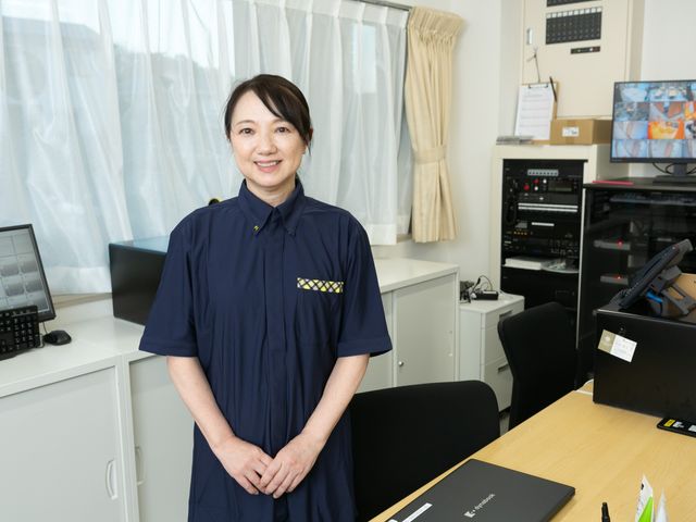 見守り室のスタッフ スタッフイメージ   見守り用のモニターや機器が整った空間で、スタッフの方が笑顔で立っています。落ち着いて働ける環境をイメージしやすいです。